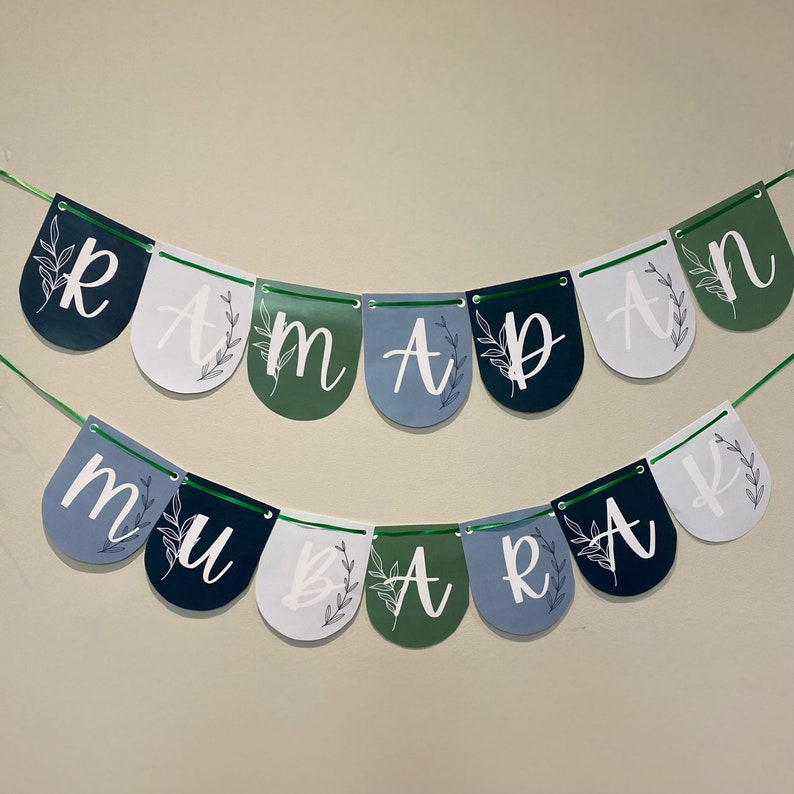 May include: A banner with the word "RAMADAN MUBARAK" spelled out on a string of colorful pennants. The pennants are in shades of blue, green, and white, with floral accents and white lettering. The banner is designed for festive occasions.