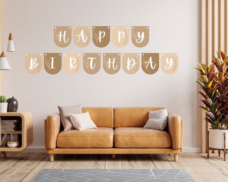 May include: A 'Happy Birthday' banner in neutral tones is suspended above a brown leather sofa. The banner comprises individual pennants in beige and brown, each displaying a white script letter. Decorative leaf motifs are also included in the design.