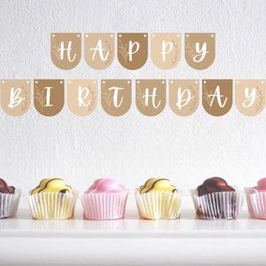 May include: A 'HAPPY BIRTHDAY' banner in white script on brown pennants with floral details. Below, six cupcakes in white paper cases with chocolate, yellow, and pink frosting are arranged on a white surface.