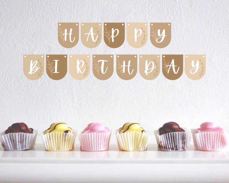 May include: A 'HAPPY BIRTHDAY' banner in white script on brown pennants with floral details. Below, six cupcakes in white paper cases with chocolate, yellow, and pink frosting are arranged on a white surface.