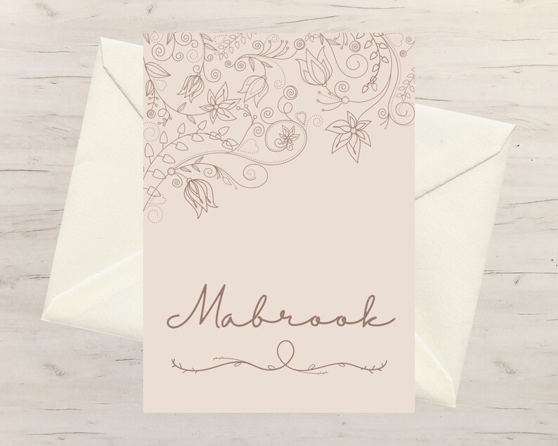 以下が含まれることがあります： ベージュ色のカードに、フローラルの線画と筆記体の「Mabrook」という名前が描かれています。カードはクリーム色の封筒の前に置かれています。フローラルデザインはカードの上部にあります。