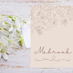以下が含まれることがあります： ベージュ色のカード。フローラルデザインと筆記体の「Mabrook」の文字。白い木製の表面に、左側に緑色の茎を持つ白い花が配置されています。
