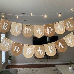 May include: A decorative banner spelling "RAMADAN MUBARAK" in white script on arched pennants. The pennants are in shades of tan and brown, with gold trim and leaf accents. A string of fairy lights hangs above the banner.