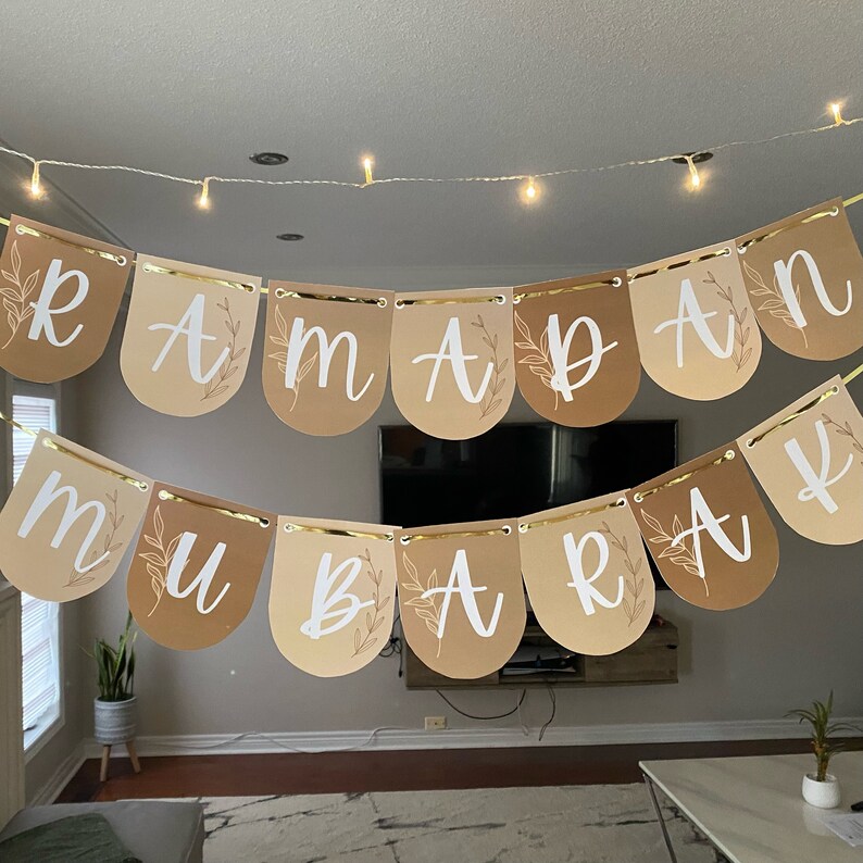 May include: A decorative banner spelling "RAMADAN MUBARAK" in white script on arched pennants. The pennants are in shades of tan and brown, with gold trim and leaf accents. A string of fairy lights hangs above the banner.