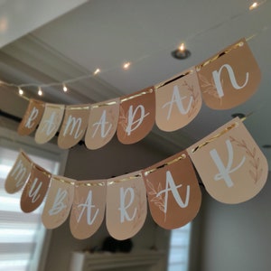 May include: A banner with the words "Ramadan Mubarak" in white script, strung across a room. The banner has a gold trim and is made of teardrop-shaped flags in shades of beige and brown. A string of fairy lights is above the banner.