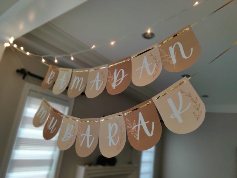 May include: A banner with the words "Ramadan Mubarak" in white script, strung across a room. The banner has a gold trim and is made of teardrop-shaped flags in shades of beige and brown. A string of fairy lights is above the banner.