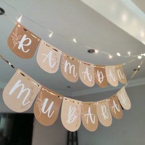 May include: A banner with the words "Ramadan Mubarak" in white script, strung across a room. The banner is made of teardrop-shaped flags in shades of tan and beige, with gold trim and floral accents. String lights are in the background.