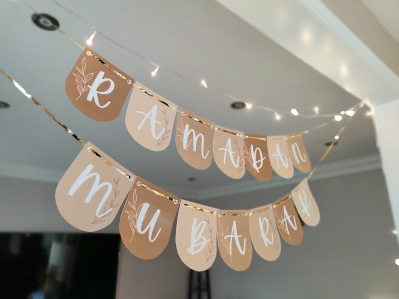May include: A banner with the words "Ramadan Mubarak" in white script, strung across a room. The banner is made of teardrop-shaped flags in shades of tan and beige, with gold trim and floral accents. String lights are in the background.