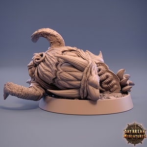 May include: A detailed, 3D printed miniature of a monstrous creature's head, lying on its side. The head is sculpted with intricate details, including sharp teeth, horns, and a textured skin. The miniature is painted in a light brown color and is set on a circular base.