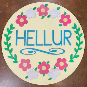 May include: A round wooden sign with a light yellow background and a blue floral wreath. The sign says "HELLUR" in blue letters.