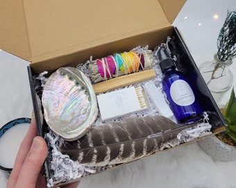 May include: A gift box filled with items for a spiritual cleansing ritual. The box contains a white sage smudge stick, a feather, a seashell, matches, and a bottle of cleansing spray. The box is lined with white tissue paper.