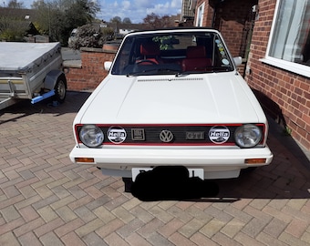 May include: White Volkswagen Golf GTI convertible with red accents and black Hella headlights. The car has a black grille with the GTI logo and a black license plate.