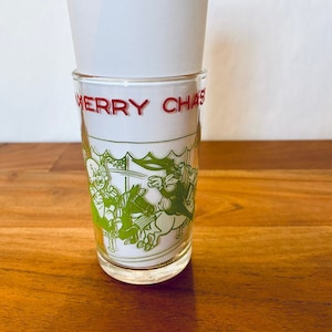 May include: A clear glass with a green and white design of cartoon characters riding a carousel. The glass has the words "Merry Christmas" in red lettering.