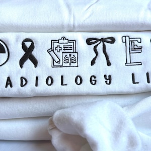 May include: A pile of white sweatshirts featuring black embroidered text and symbols. The words "RADIOLOGY LIFE" are displayed with icons including a radiation symbol, a ribbon, a medical chart, a bow tie, an X-ray machine, and a heart.