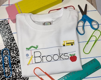 May include: A white t-shirt featuring an embroidered design of the name Brooks in black lettering on lined paper. The design includes a green caterpillar, a yellow school bus, a yellow pencil, and a red apple. The shirt is surrounded by school supplies.