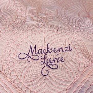 Op de afbeelding: Een roze gestikte deken met een decoratief patroon en de naam "Mackenzi Layne" geborduurd in paarse draad.
