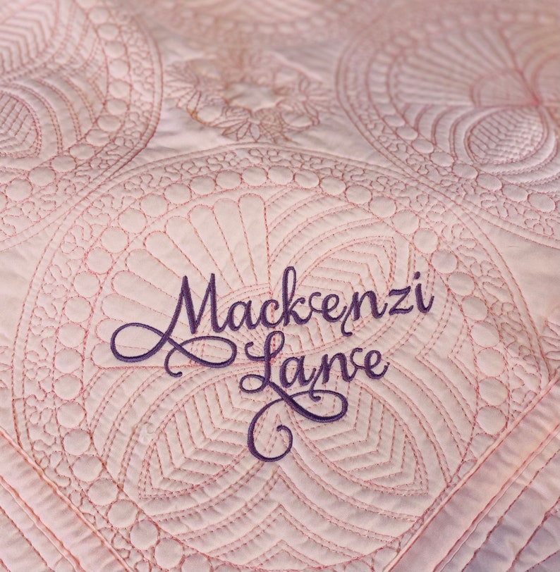 Op de afbeelding: Een roze gestikte deken met een decoratief patroon en de naam "Mackenzi Layne" geborduurd in paarse draad.