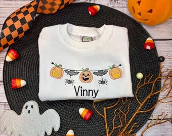 Puede incluir: Una sudadera blanca de manga larga con un diseño bordado de guirnalda de Halloween que muestra tres calabazas naranjas y dos arañas negras colgando de una cuerda. El nombre Vinny está bordado en texto negro bajo el diseño de la guirnalda.