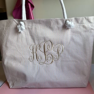 May include: A beige and white striped tote bag with white rope handles. The bag features an embroidered monogram in a gold color. The bag is made of a seersucker fabric.