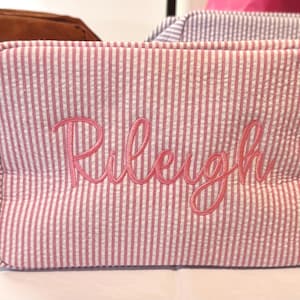 Könnte beinhalten: Kosmetiktasche mit rosa und weißen Streifen, bestickt mit dem Namen "Rileigh" in rosa Schrift. Die Tasche hat eine rechteckige Form mit Reißverschluss und kleinem Griff. Weitere Taschen sind im Hintergrund zu sehen.