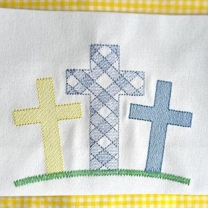 May include: Three embroidered crosses on a white fabric background. The crosses are yellow, blue, and a patterned blue and white. The crosses are on a green grass background.