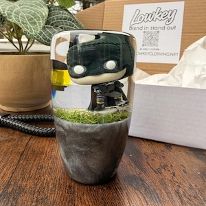 May include: A clear resin tumbler featuring a Batman Funko Pop figure. The tumbler has a gray and silver base with a patch of green faux grass. The text "lowkey" and "Blend in stand out" are visible on a box in the background.
