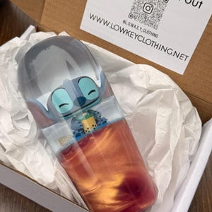 Könnte beinhalten: Ein durchsichtiger Becher mit einem Stitch-Charakter-Design, gefüllt mit einer rot-orangen Flüssigkeit. Der Becher befindet sich in einer Pappschachtel mit weißem Seidenpapier. Die Schachtel enthält auch eine weiße Karte mit dem Text "Blend in stand out" und eine Website-Adresse.