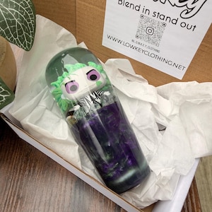 May include: A cylindrical tumbler with a Beetlejuice character design, encased in clear, green-tinted resin. The tumbler has a swirling purple and green base, and is presented in a cardboard box with the text "Lowkey Blend in stand out".