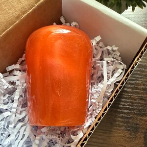 May include: A smooth, dome-shaped object with a vibrant orange, semi-translucent finish and a subtle pearlescent shimmer. The object is nestled within a cardboard box filled with white shredded paper filler, providing a secure and cushioned presentation.