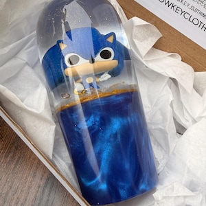 Pode incluir: Uma peça de arte cilíndrica em resina com uma figura azul de Sonic the Hedgehog. A metade inferior é preenchida com um líquido azul profundo e turbilhonante com detalhes dourados. Apresentado numa caixa forrada a branco.