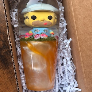 Puede incluir: Un vaso de resina transparente con una figura de personaje amarillo que lleva un sombrero azul claro y una camisa floral. La base del vaso está llena de un líquido de color ámbar arremolinado y una capa de hierba verde con pequeñas flores rosas.