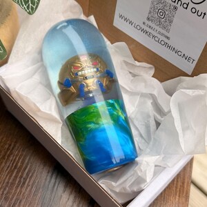 Puede incluir: Una pieza de arte de resina transparente en forma de cápsula, con una figura dorada con detalles azules, sobre un fondo azul y verde. La pieza se presenta en una caja blanca con papel de seda.