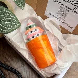 May include: A novelty sushi-themed item inside a clear, cylindrical container, presented in a white box with tissue paper. The sushi features colorful details, including pink, white, and green elements. The box also contains a tag with the text "Lowkey" and a website address.