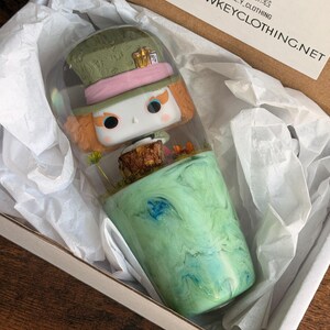 May include: A Mad Hatter Funko Pop figure inside a clear container, set in a green and blue resin base. The figure is in a cardboard box with white tissue paper. The box also has a QR code and the website address: WWW.LOWKEYCLOTHING.NET.