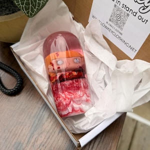 Puede incluir: Una pieza de arte decorativa en capas en una caja. La pieza presenta una serie de figuras tipo muñeca encerradas en una forma cilíndrica transparente. Las figuras tienen tonos naranja y rojo, con una base veteada roja y blanca. La caja contiene el texto "Blend in stand out".