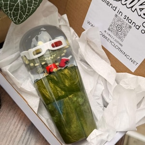 以下が含まれることがあります： 3Dメガネをかけた漫画のアリゲーターが飲み物を持っている、手作りの樹脂タンブラー。タンブラーは緑と金のマーブル模様で、パッケージには「Blend in stand out」の文字が見える。タンブラーは箱に入っている。