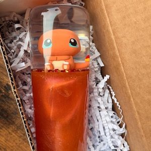 May include: A clear cylindrical tumbler featuring a small orange Charmander figurine inside the top section. The lower portion of the tumbler is filled with a vibrant, shimmering orange liquid, separated from the figurine by a layer of red glitter.