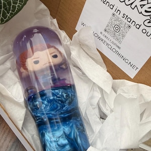 May include: A custom-made tumbler with a Funko Pop figure inside, featuring a swirling blue and purple design. The tumbler is in a cardboard box with tissue paper and a white label that says "Lowkey Clothing".