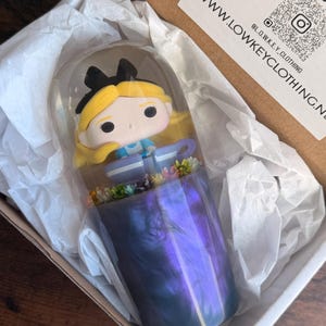 以下が含まれることがあります： 不思議の国のアリスのフィギュアが入った装飾ガラスドーム。ベースは渦巻く紫と青の樹脂です。フィギュアは小さな花に囲まれています。商品は白いティッシュペーパーが入った段ボール箱に梱包されています。