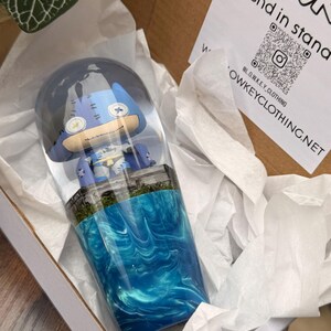 Peut inclure: Un gobelet bleu et blanc avec un personnage de dessin animé à l'intérieur d'un dôme en résine transparente. Le gobelet présente un motif d'eau tourbillonnante bleue et blanche. Il est dans une boîte en carton avec du papier de soie blanc. Le texte "Blend in stand" et un QR code sont visibles.
