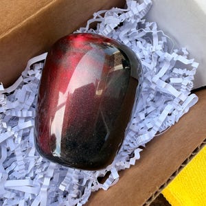 May include: A smooth, rounded object with a glossy finish featuring a gradient of deep red and black colors with subtle embedded glitter particles. The object has a reflective surface and is nestled within a cardboard box filled with white paper shreds.