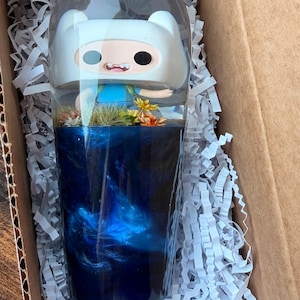 Puede incluir: Un vaso transparente que presenta una figura de Finn el Humano incrustada en la parte superior, con una base llena de resina arremolinada en azul oscuro y negro. La sección central contiene pequeños elementos florales artificiales verdes y amarillos.