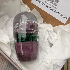 May include: A clear, cylindrical container filled with purple glitter, green dice, and a purple, speckled object. The container is inside a white box with tissue paper. The website address www.LOWKEYCLOTHING.NET is visible.
