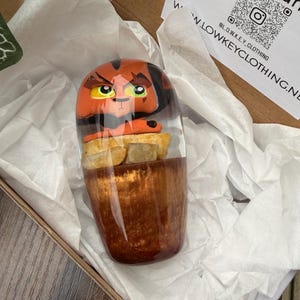 Pode incluir: Um copo decorativo com um design de personagem de desenho animado em laranja, preto e amarelo. O copo é apresentado em uma caixa de papelão com papel de seda branco. O copo tem uma base marrom e um topo transparente, mostrando o rosto do personagem.