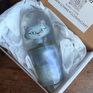 Op de afbeelding: Een grillige kattenfiguur in een heldere, klokvormige behuizing, geplaatst op een parelmoer, iriserende basis. De kat heeft een lichtblauw lichaam met turquoise strepen en grote, ronde ogen. De figuur wordt gepresenteerd in een kartonnen doos met vloeipapier.