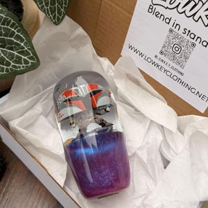 May include: A clear resin tumbler with a design featuring a character in a red and gray helmet. The bottom half of the tumbler is a mix of purple and blue glitter. The tumbler is inside a cardboard box with white tissue paper.