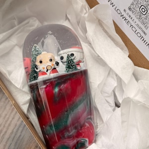 以下が含まれることがあります： サンタ帽をかぶった2体のフィギュアと小さなクリスマスツリーが入ったスノードーム。ベースは赤、緑、白の渦巻き模様。スノードームは、白い薄葉紙が入った段ボール箱に入っています。