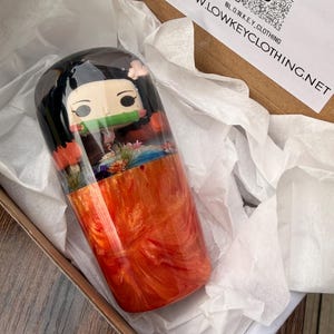 Op de afbeelding: Een harsbeker met een karakterontwerp met zwart haar, een groene mondbeschermer en een roze bloem. De beker heeft een oranje en gouden wervelontwerp en is verpakt in een kartonnen doos met wit vloeipapier.