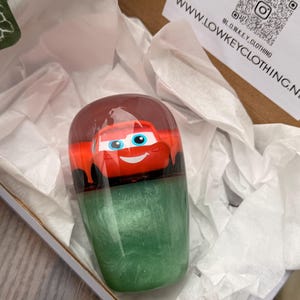 May include: A resin tumbler featuring a red cartoon car design on top, with a green glittery base. The tumbler is presented inside a cardboard box with white tissue paper. The website address www.LOWKEYCLOTHING.NZ is visible on a white card.