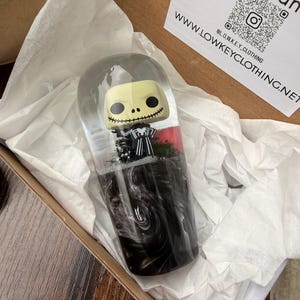May include: A clear, capsule-shaped resin art piece featuring a Jack Skellington figure. The figure is set against a backdrop of miniature trees and red accents, with swirling black resin at the base. The piece is inside a cardboard box.